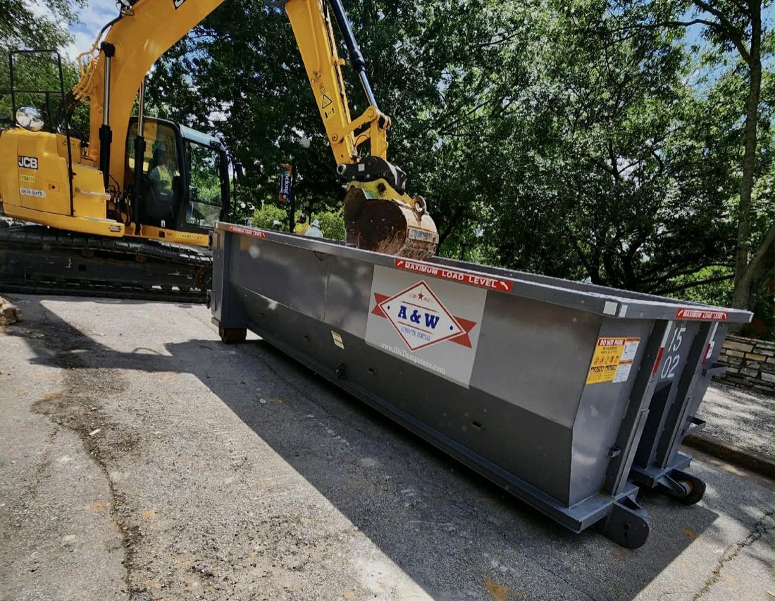 Excavator loading an A&W branded roll-off dumpster on a demolition job